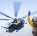 A military helicopter, appearing to be a CH-53E Super Stallion, is captured in a low-angle shot during a deck landing or departure operation. A flight deck crew member in yellow jersey and white helmet is giving hand signals to the aircraft. The rotor blades show significant motion blur, indicating active rotation at high speed.