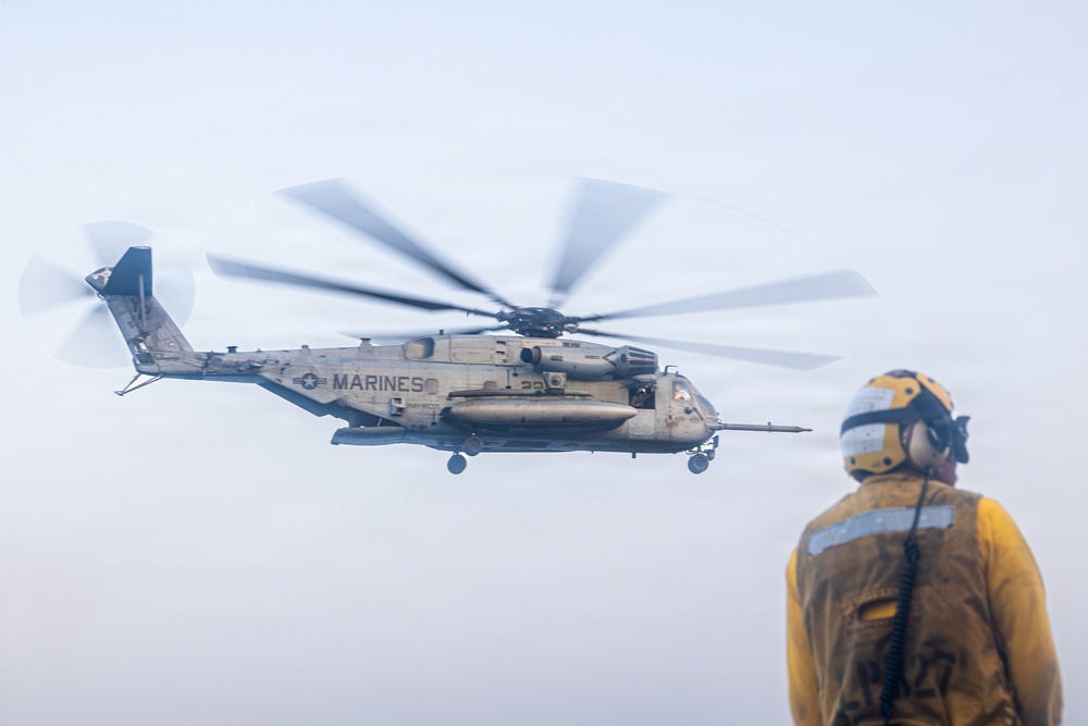 A U.S. Marines heavy-lift helicopter, identifiable as a CH-53E Super Stallion, is hovering or maneuvering in hazy/overcast conditions. A flight deck crew member wearing yellow protective gear, a white helmet, and hearing protection stands in the foreground observing the aircraft. The scene appears to be taking place on a naval vessel's flight deck.
