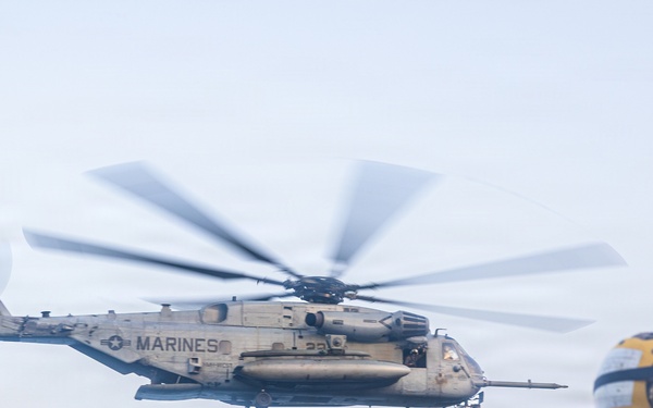 A U.S. Marines heavy-lift helicopter, identifiable as a CH-53E Super Stallion, is hovering or maneuvering in hazy/overcast conditions. A flight deck crew member wearing yellow protective gear, a white helmet, and hearing protection stands in the foreground observing the aircraft. The scene appears to be taking place on a naval vessel's flight deck.