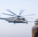 A U.S. Marines heavy-lift helicopter, identifiable as a CH-53E Super Stallion, is hovering or maneuvering in hazy/overcast conditions. A flight deck crew member wearing yellow protective gear, a white helmet, and hearing protection stands in the foreground observing the aircraft. The scene appears to be taking place on a naval vessel's flight deck.