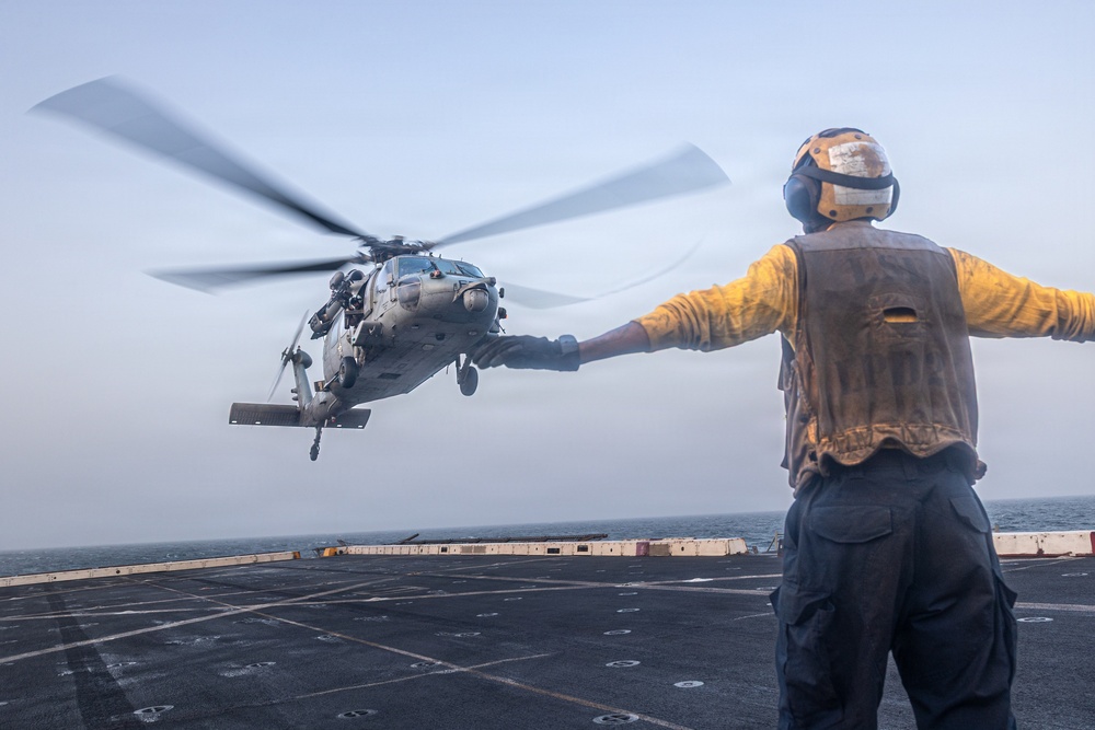 A naval flight deck crew member in a yellow shirt and safety vest directs a military helicopter during landing or takeoff operations on a ship's flight deck at sea. The handler is using standard hand signals with arms outstretched to guide the aircraft. The helicopter, identifiable as a Sikorsky MH-60 Seahawk variant, is hovering just above the deck with rotor blades in motion.