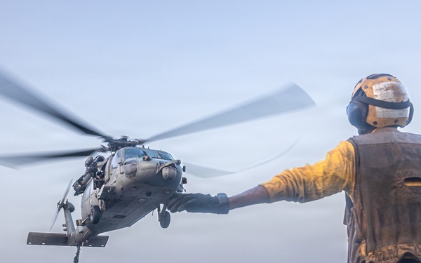 A naval flight deck crew member in a yellow shirt and safety vest directs a military helicopter during landing or takeoff operations on a ship's flight deck at sea. The handler is using standard hand signals with arms outstretched to guide the aircraft. The helicopter, identifiable as a Sikorsky MH-60 Seahawk variant, is hovering just above the deck with rotor blades in motion.
