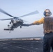 A naval flight deck crew member in a yellow shirt and safety vest directs a military helicopter during landing or takeoff operations on a ship's flight deck at sea. The handler is using standard hand signals with arms outstretched to guide the aircraft. The helicopter, identifiable as a Sikorsky MH-60 Seahawk variant, is hovering just above the deck with rotor blades in motion.