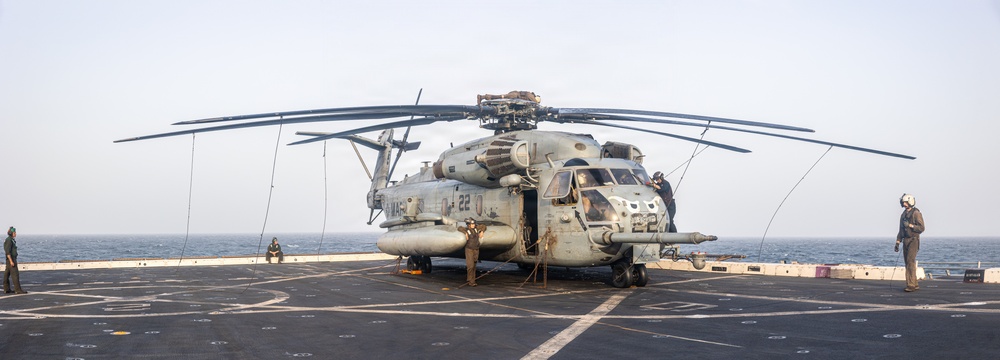 A large military helicopter, identified as a CH-53E Super Stallion bearing the number '22' and markings consistent with the U.S. Marine Corps, is secured on the flight deck of a naval vessel at sea. Multiple personnel in flight deck gear are positioned around the aircraft, appearing to conduct maintenance or tie-down operations. The ocean horizon is visible in the background under an overcast sky.