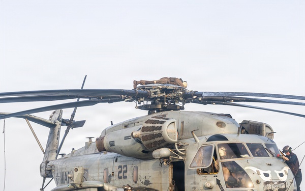 A large military helicopter, identified as a CH-53E Super Stallion bearing the number '22' and markings consistent with the U.S. Marine Corps, is secured on the flight deck of a naval vessel at sea. Multiple personnel in flight deck gear are positioned around the aircraft, appearing to conduct maintenance or tie-down operations. The ocean horizon is visible in the background under an overcast sky.