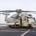 A large military helicopter, identified as a CH-53E Super Stallion bearing the number '22' and markings consistent with the U.S. Marine Corps, is secured on the flight deck of a naval vessel at sea. Multiple personnel in flight deck gear are positioned around the aircraft, appearing to conduct maintenance or tie-down operations. The ocean horizon is visible in the background under an overcast sky.