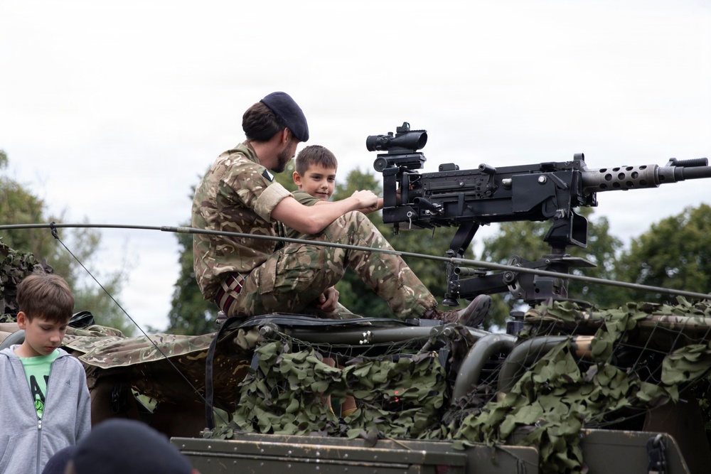 A military personnel member in British DPM camouflage uniform and dark beret is showing a young boy a mounted machine gun on top of a military vehicle covered in camouflage netting. The setting appears to be a public military display or open day event. At least two other children are visible nearby observing the vehicle.