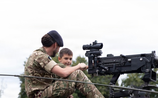 A military personnel member in British DPM camouflage uniform and dark beret is showing a young boy a mounted machine gun on top of a military vehicle covered in camouflage netting. The setting appears to be a public military display or open day event. At least two other children are visible nearby observing the vehicle.