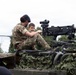 A military personnel member in British DPM camouflage uniform and dark beret is showing a young boy a mounted machine gun on top of a military vehicle covered in camouflage netting. The setting appears to be a public military display or open day event. At least two other children are visible nearby observing the vehicle.
