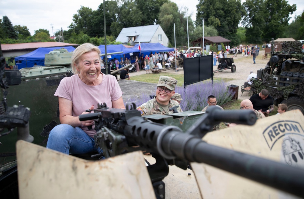A civilian woman in a pink shirt is smiling broadly while handling a mounted machine gun on a military vehicle at what appears to be a public military exhibition or open day event. A U.S. Army soldier with the name tape 'Williams' is visible beside her, also smiling. The background shows a crowded outdoor venue with military equipment displays, blue tents, and numerous visitors.