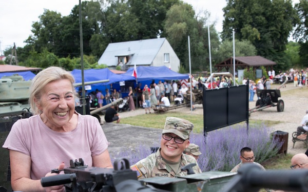A civilian woman in a pink shirt is smiling broadly while handling a mounted machine gun on a military vehicle at what appears to be a public military exhibition or open day event. A U.S. Army soldier with the name tape 'Williams' is visible beside her, also smiling. The background shows a crowded outdoor venue with military equipment displays, blue tents, and numerous visitors.