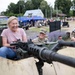 A civilian woman in a pink shirt is smiling broadly while handling a mounted machine gun on a military vehicle at what appears to be a public military exhibition or open day event. A U.S. Army soldier with the name tape 'Williams' is visible beside her, also smiling. The background shows a crowded outdoor venue with military equipment displays, blue tents, and numerous visitors.