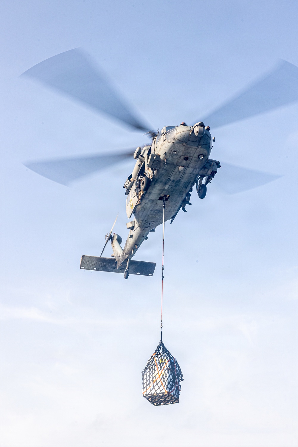 A military helicopter is captured in flight from a low-angle perspective, carrying a cargo net sling load suspended beneath it. The helicopter appears to be a large, multi-blade naval or military transport variant performing a vertical replenishment or cargo lift operation. The rotor blades show motion blur, indicating active flight.