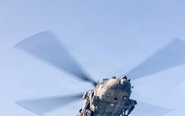 A military helicopter is captured in flight from a low-angle perspective, carrying a cargo net sling load suspended beneath it. The helicopter appears to be a large, multi-blade naval or military transport variant performing a vertical replenishment or cargo lift operation. The rotor blades show motion blur, indicating active flight.