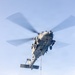 A military helicopter is captured in flight from a low-angle perspective, carrying a cargo net sling load suspended beneath it. The helicopter appears to be a large, multi-blade naval or military transport variant performing a vertical replenishment or cargo lift operation. The rotor blades show motion blur, indicating active flight.