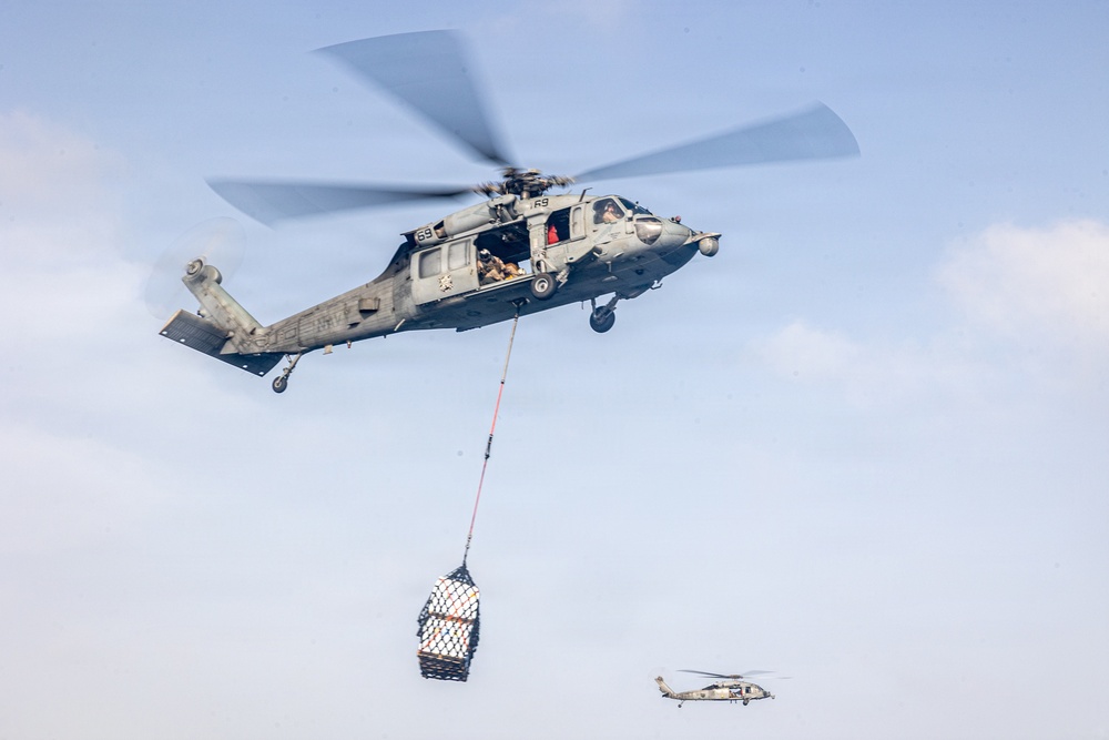 A military helicopter, identified as number 69, is performing a vertical replenishment (VERTREP) operation, carrying a cargo net loaded with supplies suspended beneath it via a cable. A second similar helicopter is visible in the background at a lower altitude. The scene takes place over what appears to be an open sky with light cloud cover.