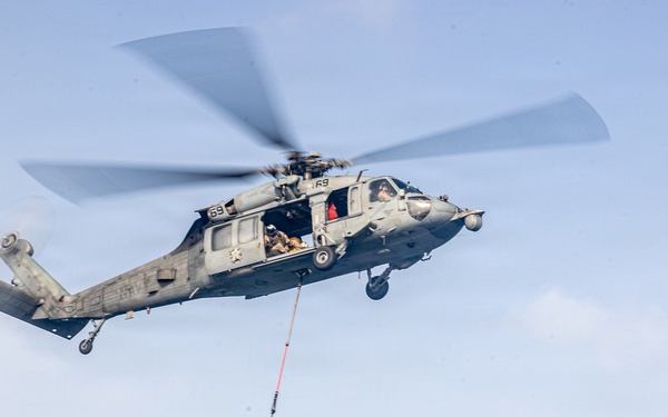 A military helicopter, identified as number 69, is performing a vertical replenishment (VERTREP) operation, carrying a cargo net loaded with supplies suspended beneath it via a cable. A second similar helicopter is visible in the background at a lower altitude. The scene takes place over what appears to be an open sky with light cloud cover.