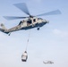 A military helicopter, identified as number 69, is performing a vertical replenishment (VERTREP) operation, carrying a cargo net loaded with supplies suspended beneath it via a cable. A second similar helicopter is visible in the background at a lower altitude. The scene takes place over what appears to be an open sky with light cloud cover.