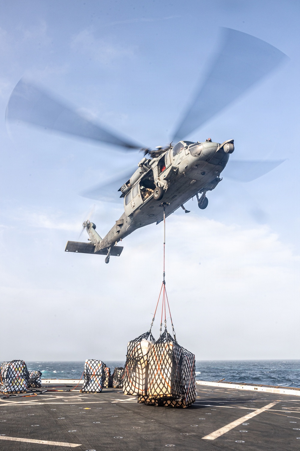 A military helicopter, appearing to be a U.S. Navy MH-60 Seahawk, is conducting a vertical replenishment (VERTREP) operation over the flight deck of a naval vessel at sea. The helicopter is airborne and lifting a cargo net containing supplies, with additional netted cargo pallets visible on the deck below. The operation is taking place in open ocean conditions under a partly cloudy sky.