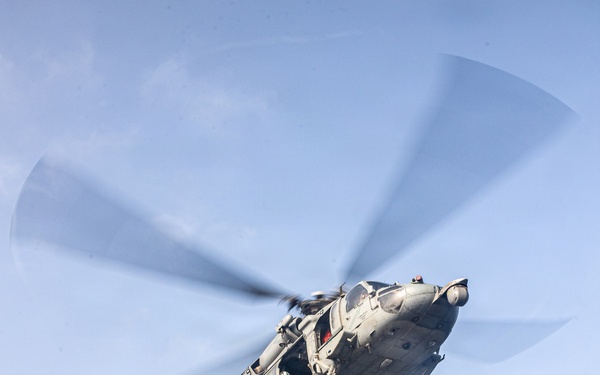 A military helicopter, appearing to be a U.S. Navy MH-60 Seahawk, is conducting a vertical replenishment (VERTREP) operation over the flight deck of a naval vessel at sea. The helicopter is airborne and lifting a cargo net containing supplies, with additional netted cargo pallets visible on the deck below. The operation is taking place in open ocean conditions under a partly cloudy sky.