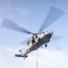 A military helicopter, appearing to be a U.S. Navy MH-60 Seahawk, is conducting a vertical replenishment (VERTREP) operation over the flight deck of a naval vessel at sea. The helicopter is airborne and lifting a cargo net containing supplies, with additional netted cargo pallets visible on the deck below. The operation is taking place in open ocean conditions under a partly cloudy sky.