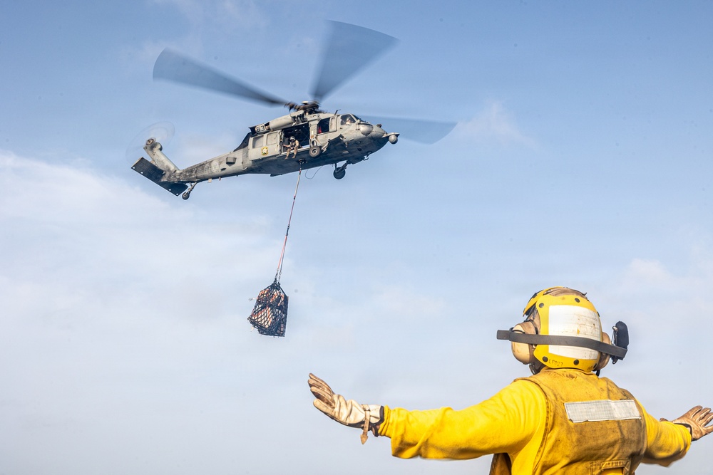 A military helicopter, likely a Sikorsky MH-60 Seahawk, hovers above a naval vessel conducting a vertical replenishment (VERTREP) operation. A deck crew member dressed in yellow flight deck gear is directing the helicopter with outstretched arms as a cargo net loaded with supplies is suspended below the aircraft. This appears to be a standard underway replenishment operation conducted by a naval aviation unit.