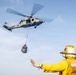 A military helicopter, likely a Sikorsky MH-60 Seahawk, hovers above a naval vessel conducting a vertical replenishment (VERTREP) operation. A deck crew member dressed in yellow flight deck gear is directing the helicopter with outstretched arms as a cargo net loaded with supplies is suspended below the aircraft. This appears to be a standard underway replenishment operation conducted by a naval aviation unit.