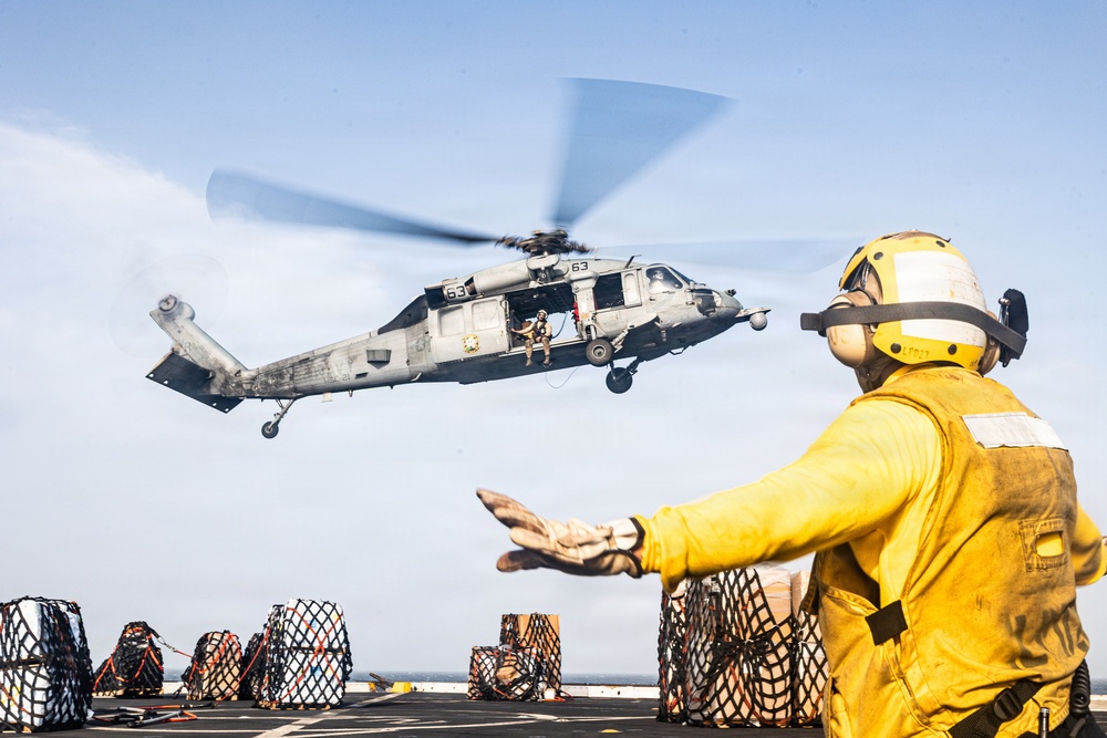 A U.S. Navy helicopter, marked with the number '63', hovers over the flight deck of a naval vessel during what appears to be a vertical replenishment (VERTREP) operation. A deck crew member in yellow safety gear and helmet is directing the helicopter with hand signals. Multiple cargo pallets secured with netting are visible on the deck, indicating an active logistics or resupply mission.