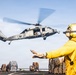 A U.S. Navy helicopter, marked with the number '63', hovers over the flight deck of a naval vessel during what appears to be a vertical replenishment (VERTREP) operation. A deck crew member in yellow safety gear and helmet is directing the helicopter with hand signals. Multiple cargo pallets secured with netting are visible on the deck, indicating an active logistics or resupply mission.