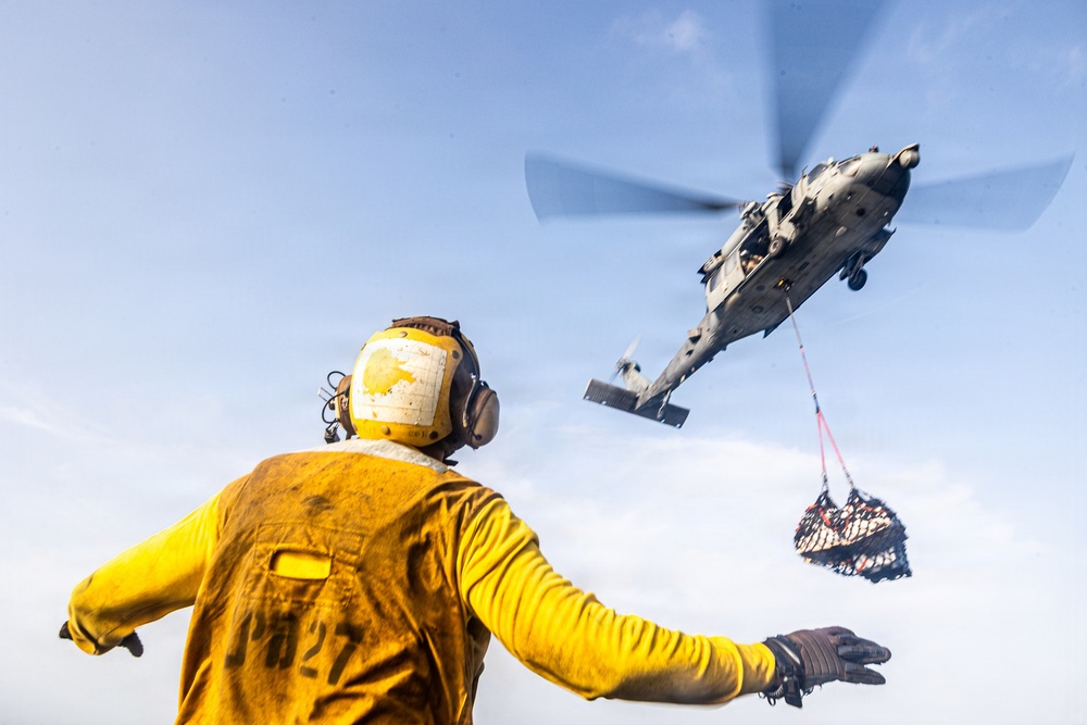 A naval deck crew member wearing a yellow jersey and flight deck helmet with ear protection is directing or signaling a military helicopter performing a vertical replenishment (VERTREP) operation. The helicopter, appearing to be an MH-60 Seahawk variant, is airborne and carrying a cargo net loaded with supplies suspended by a sling line. This type of operation is commonly used to transfer supplies between ships at sea.