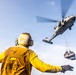 A naval deck crew member wearing a yellow jersey and flight deck helmet with ear protection is directing or signaling a military helicopter performing a vertical replenishment (VERTREP) operation. The helicopter, appearing to be an MH-60 Seahawk variant, is airborne and carrying a cargo net loaded with supplies suspended by a sling line. This type of operation is commonly used to transfer supplies between ships at sea.