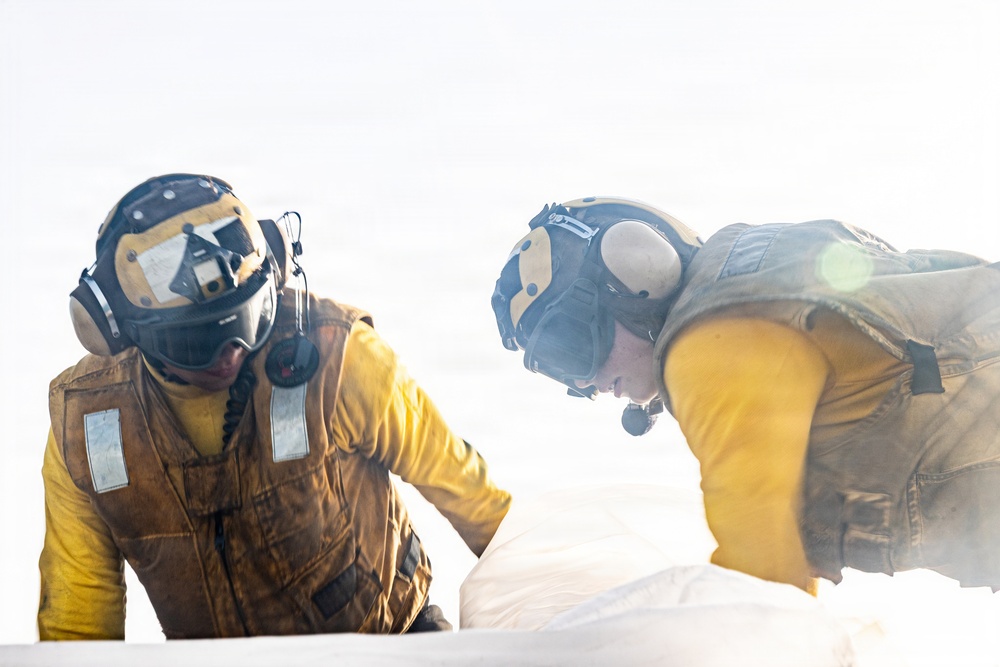 Two personnel wearing yellow flight deck jerseys and protective helmets with ear defenders are leaning over and working on a white surface, likely an aircraft on a naval flight deck. Both individuals are equipped with communication headsets and protective gear consistent with carrier deck crew operations. The image is slightly hazy with lens flare, suggesting bright outdoor lighting conditions.