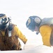 Two personnel wearing yellow flight deck jerseys and protective helmets with ear defenders are leaning over and working on a white surface, likely an aircraft on a naval flight deck. Both individuals are equipped with communication headsets and protective gear consistent with carrier deck crew operations. The image is slightly hazy with lens flare, suggesting bright outdoor lighting conditions.