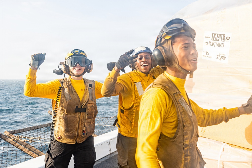 Three naval personnel in yellow flight deck jerseys and helmets with communication headsets are celebrating on the deck of a ship at sea. They appear to be reacting joyfully, with raised fists and smiles, near a large white container or mail bag labeled 'MAIL TO PORTLAND.' The image captures a candid, high-morale moment aboard what appears to be a U.S. Navy vessel.