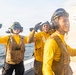 Three naval personnel in yellow flight deck jerseys and helmets with communication headsets are celebrating on the deck of a ship at sea. They appear to be reacting joyfully, with raised fists and smiles, near a large white container or mail bag labeled 'MAIL TO PORTLAND.' The image captures a candid, high-morale moment aboard what appears to be a U.S. Navy vessel.