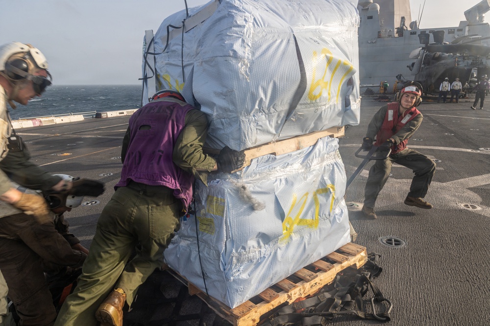 Military personnel are handling a large wrapped cargo pallet on what appears to be the flight deck of a naval vessel at sea. The crew members, wearing color-coded safety vests and helmets, are pushing and pulling the pallet across the deck. A helicopter and additional personnel are visible in the background near the ship's superstructure.