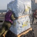Military personnel are handling a large wrapped cargo pallet on what appears to be the flight deck of a naval vessel at sea. The crew members, wearing color-coded safety vests and helmets, are pushing and pulling the pallet across the deck. A helicopter and additional personnel are visible in the background near the ship's superstructure.
