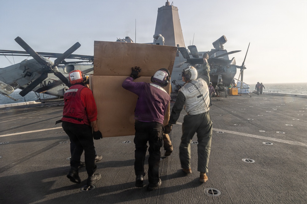 Three personnel in flight deck gear are carrying a large cardboard box or panel across the flight deck of a naval vessel. The scene appears to be on an amphibious assault ship or similar warship, with rotary-wing aircraft (likely CH-53 or MV-22 type) visible in the background. A ship's superstructure with a distinctive angular mast is visible in the mid-background.