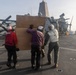 Three personnel in flight deck gear are carrying a large cardboard box or panel across the flight deck of a naval vessel. The scene appears to be on an amphibious assault ship or similar warship, with rotary-wing aircraft (likely CH-53 or MV-22 type) visible in the background. A ship's superstructure with a distinctive angular mast is visible in the mid-background.
