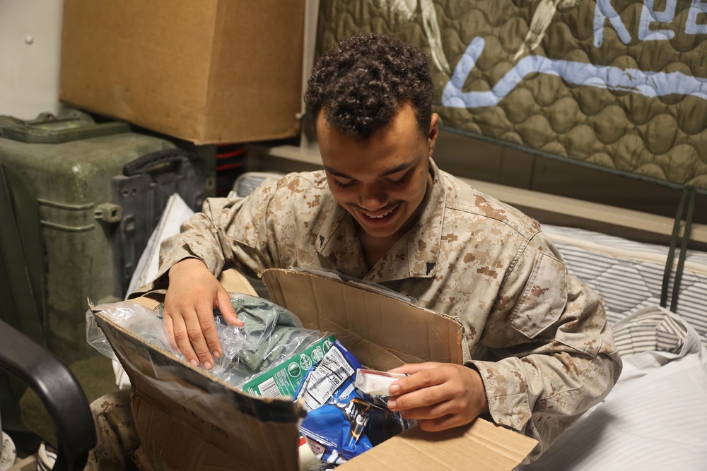 A young service member in desert MARPAT camouflage uniform is smiling while opening a care package box. The box contains various food and personal items, suggesting it was sent from home or a support organization. The setting appears to be a military barracks or billet room.