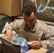 A young service member in desert MARPAT camouflage uniform is smiling while opening a care package box. The box contains various food and personal items, suggesting it was sent from home or a support organization. The setting appears to be a military barracks or billet room.