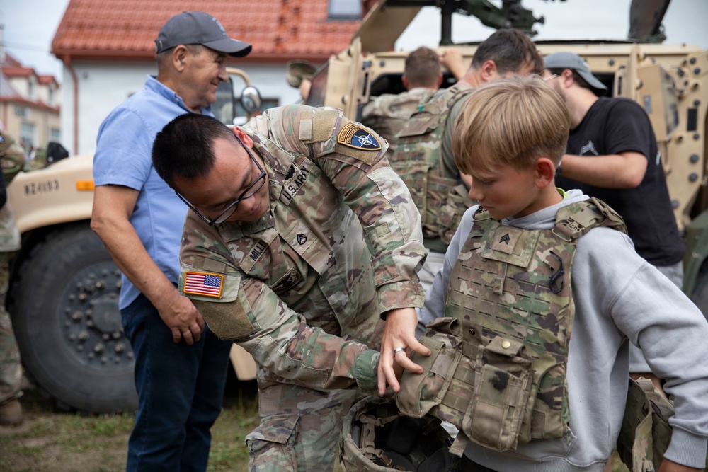 A U.S. Army soldier is helping a young boy try on a tactical body armor vest during what appears to be a public military outreach or open house event. The soldier, wearing U.S. Army and NATO patches, is bending down to adjust the equipment on the child. A military vehicle (HMMWV/Humvee) is visible in the background along with several other civilians and personnel.