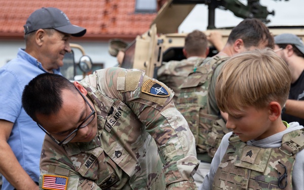 A U.S. Army soldier is helping a young boy try on a tactical body armor vest during what appears to be a public military outreach or open house event. The soldier, wearing U.S. Army and NATO patches, is bending down to adjust the equipment on the child. A military vehicle (HMMWV/Humvee) is visible in the background along with several other civilians and personnel.