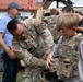 A U.S. Army soldier is helping a young boy try on a tactical body armor vest during what appears to be a public military outreach or open house event. The soldier, wearing U.S. Army and NATO patches, is bending down to adjust the equipment on the child. A military vehicle (HMMWV/Humvee) is visible in the background along with several other civilians and personnel.