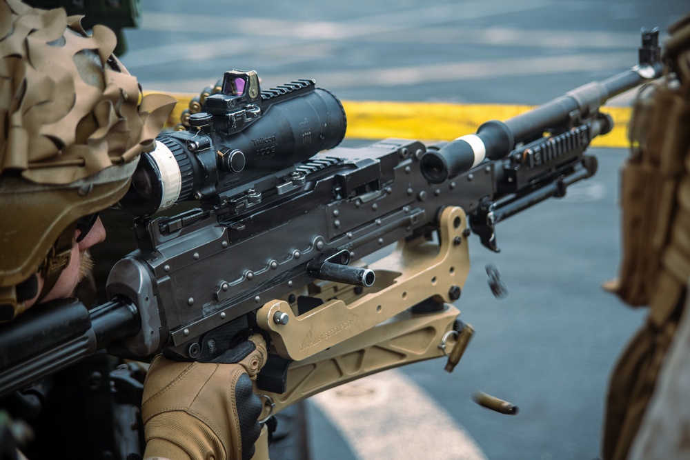 A soldier in camouflage uniform is actively firing an M240 machine gun, visible by the spent brass casings ejecting from the weapon. The firearm is equipped with a Trijicon ACOG scope with a red dot sight mounted on top. The image captures the weapon in mid-fire on what appears to be a military vessel or hardstand surface.