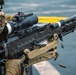 A soldier in camouflage uniform is actively firing an M240 machine gun, visible by the spent brass casings ejecting from the weapon. The firearm is equipped with a Trijicon ACOG scope with a red dot sight mounted on top. The image captures the weapon in mid-fire on what appears to be a military vessel or hardstand surface.