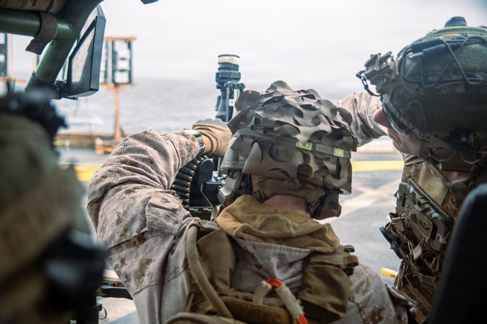 Two military personnel in combat gear are positioned on what appears to be a ship's deck, with one operating or adjusting a crew-served weapon system. Both wear helmets with camouflage covers and tactical equipment. The setting appears to be a maritime environment based on the visible ocean and ship deck infrastructure in the background.