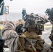 Two military personnel in combat gear are positioned on what appears to be a ship's deck, with one operating or adjusting a crew-served weapon system. Both wear helmets with camouflage covers and tactical equipment. The setting appears to be a maritime environment based on the visible ocean and ship deck infrastructure in the background.