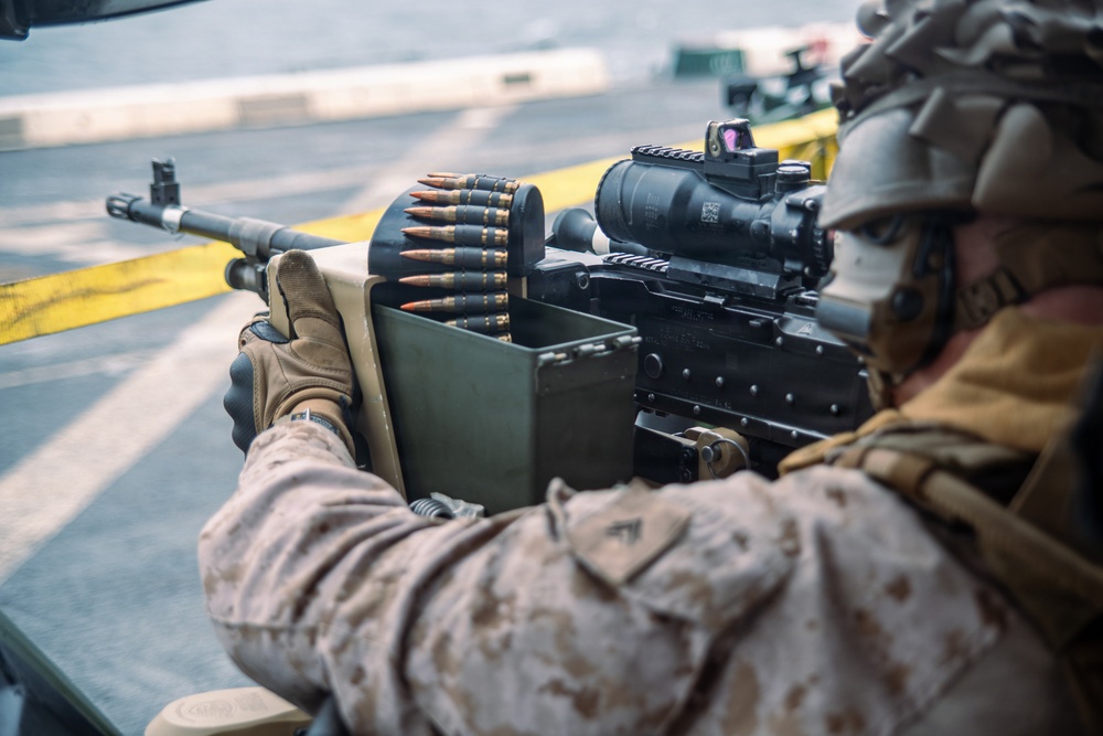 A military service member in desert MARPAT camouflage uniform is operating a belt-fed machine gun, likely an M249 SAW or M240, mounted in what appears to be a vehicle or aircraft doorway. The weapon is equipped with a magnified optic (ACOG-style) and a red dot sight mounted on top. An ammunition box with a linked belt of rounds is visible feeding into the weapon.