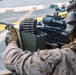 A military service member in desert MARPAT camouflage uniform is operating a belt-fed machine gun, likely an M249 SAW or M240, mounted in what appears to be a vehicle or aircraft doorway. The weapon is equipped with a magnified optic (ACOG-style) and a red dot sight mounted on top. An ammunition box with a linked belt of rounds is visible feeding into the weapon.